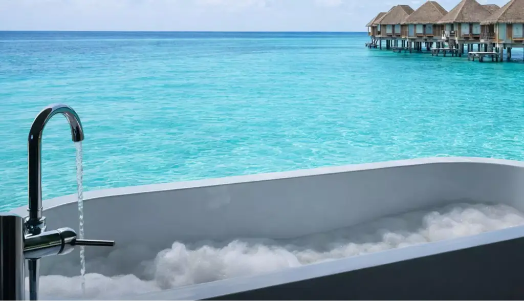 Visualización arquitectónica de una bañera de burbujas exterior frente a un mar turquesa en calma con villas sobre pilotes al fondo.