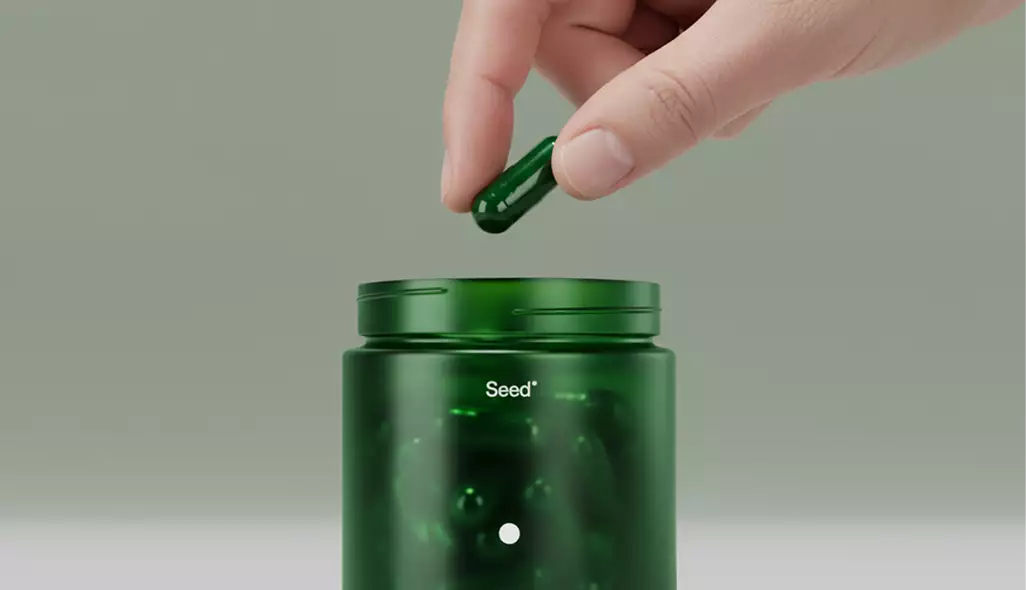 Hand laat een groene capsule vallen in een open groene pot met Seed-etiket op neutrale achtergrond, verpakkingsontwerp voor supplementen