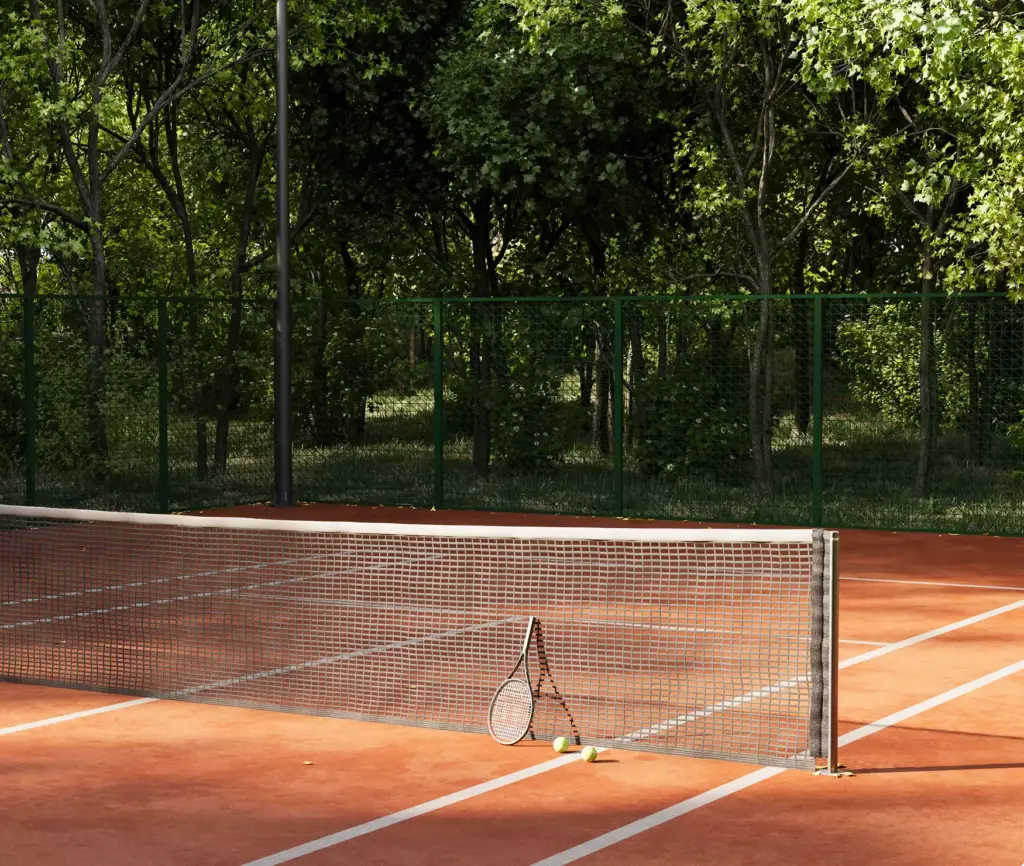 Campo da tennis in terra battuta con recinzione verde tra gli alberi, rete con racchetta e palline in piena luce.