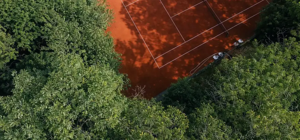 Campo da tennis in terra battuta circondato da alberi fitti, visto dall'alto con le ombre sul manto di gioco.