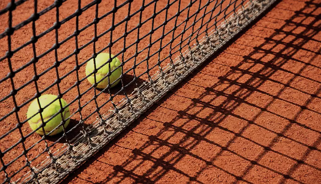 Palline da tennis accanto alla rete sul campo in terra battuta, con il disegno delle ombre sulla superficie rossa.
