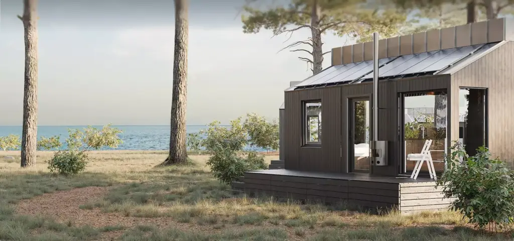 Render van een compacte prefab woning met donkere houten gevelbekleding en een schuin dak met zonnepanelen, een glazen pui naar een houten terras met een witte klapstoel, een rookkanaal langs de gevel, tussen dennen aan een strook water