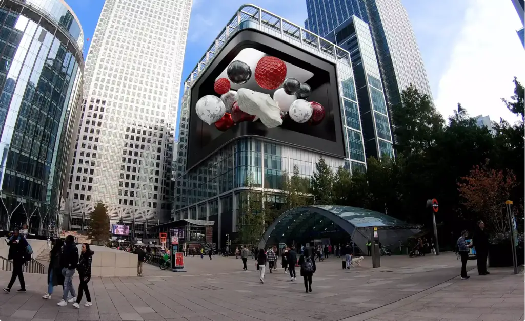 Fooh ads su un gigantesco maxischermo 3D sopra una piazza cittadina, con torri, pedoni e l'ingresso di una stazione