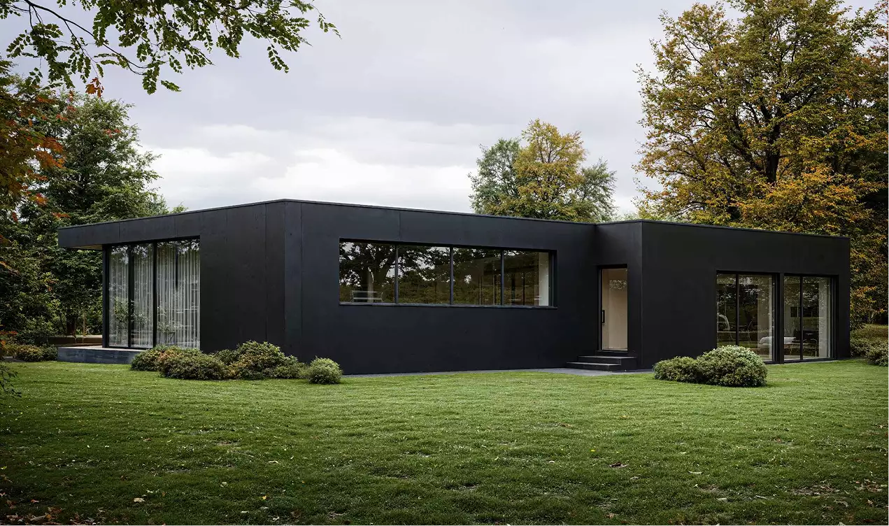 Maison de plain-pied à toit plat et bardage noir mat, longue fenêtre bandeau et baies coulissantes, posée sur une pelouse bordée d'arbres