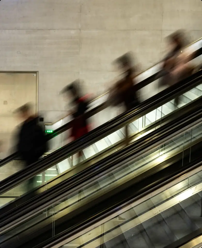 Pendolari in movimento su una scala mobile ripresi in mosso, a illustrare la vita quotidiana frenetica per cui è pensata un'app di salute digitale.