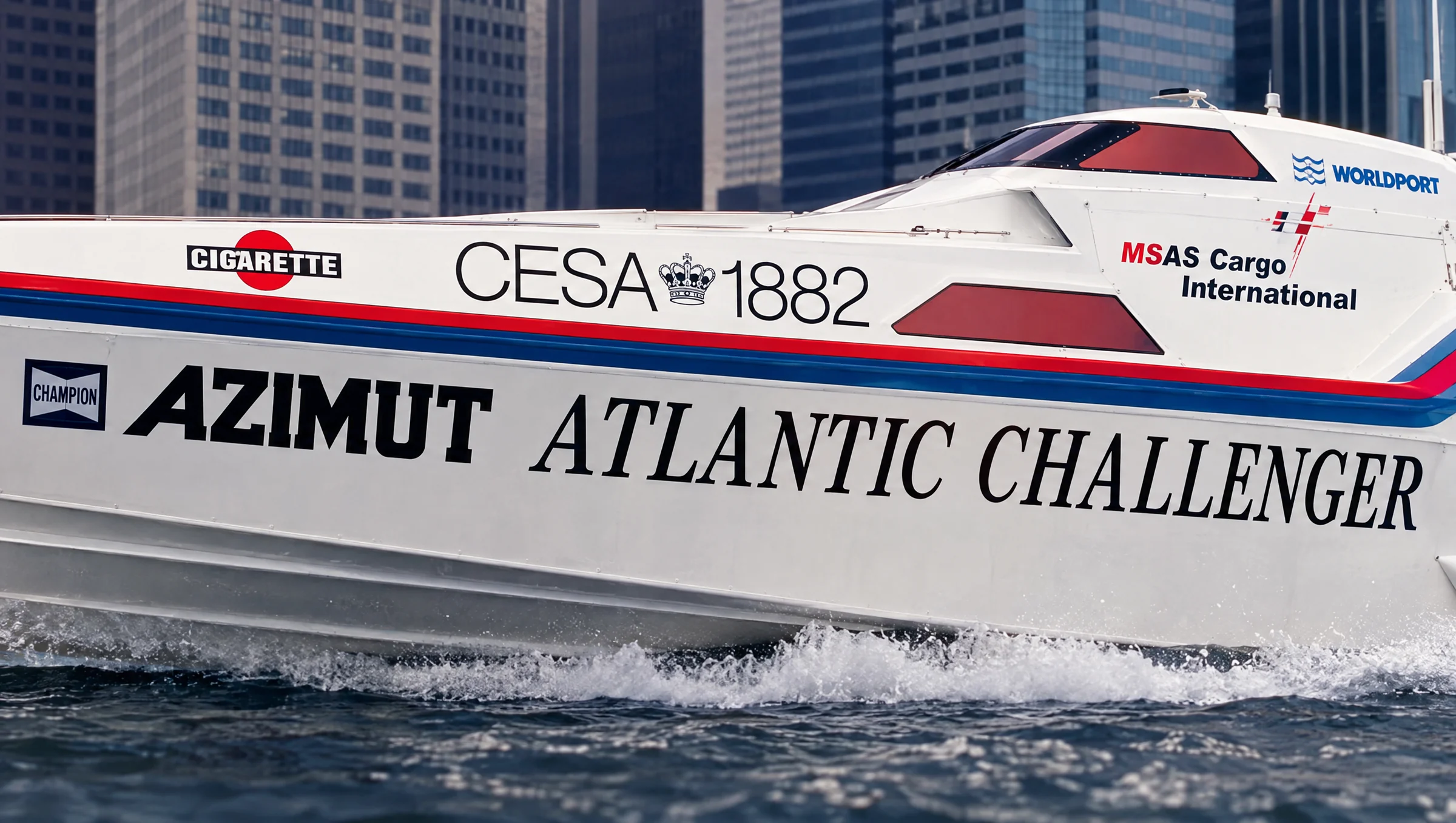 Primo piano dello scafo di un motoscafo offshore con livrea da corsa e loghi degli sponsor davanti agli alti edifici di una città