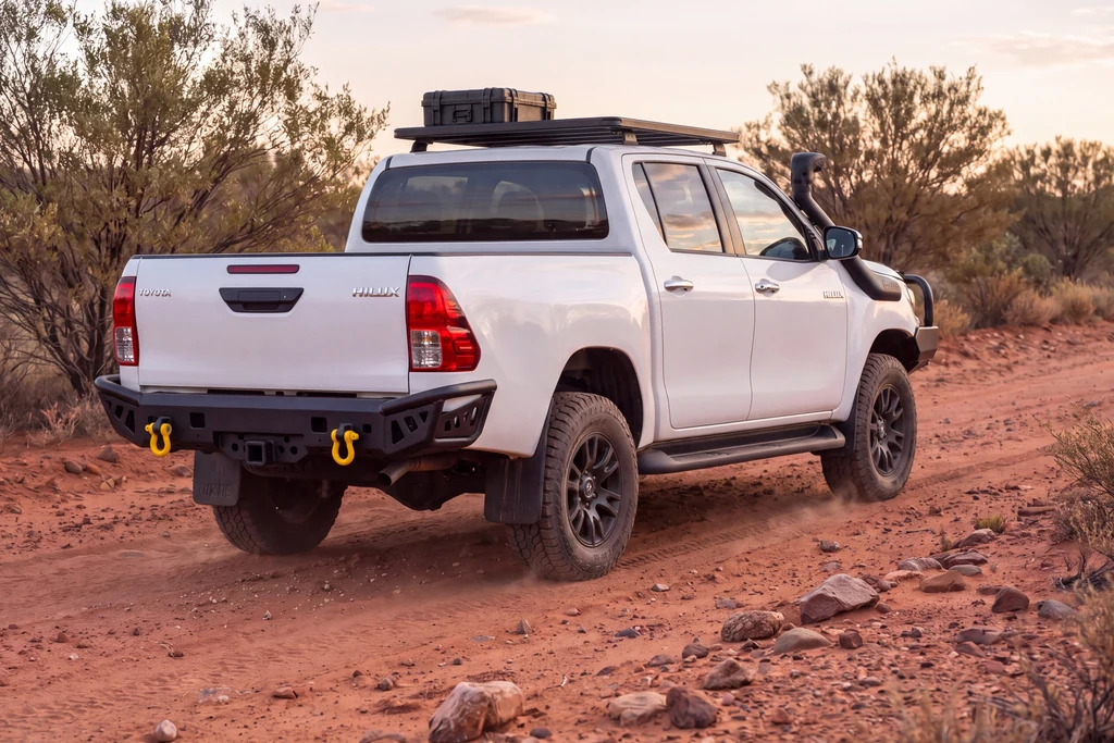 Render of a white Toyota Hilux with a roof rack and a black steel rear bar on a red dirt outback track