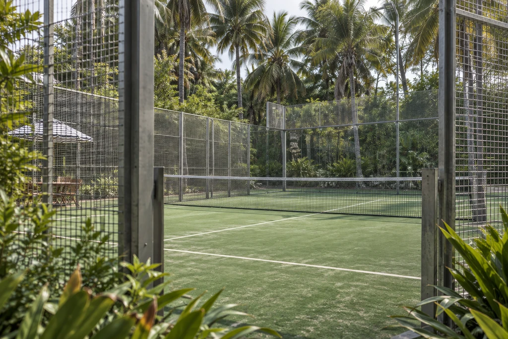 CGI auf Augenhöhe vom Eingangstor des privaten Padelplatzes mit Gitterzaun, Sonnenschirmen und tropischem Garten.