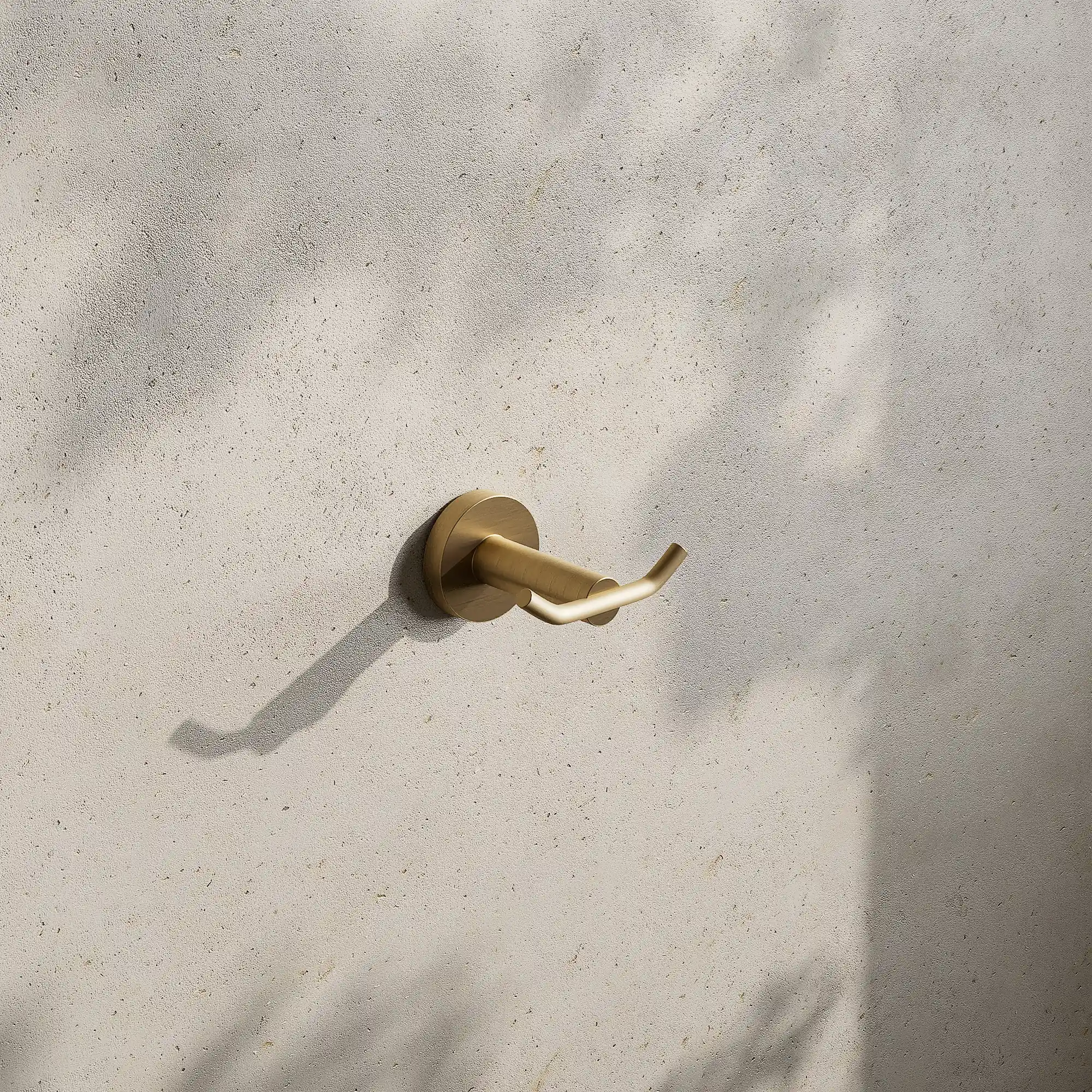 Render of a double robe hook in brushed gold on a stone wall with leaf shadows