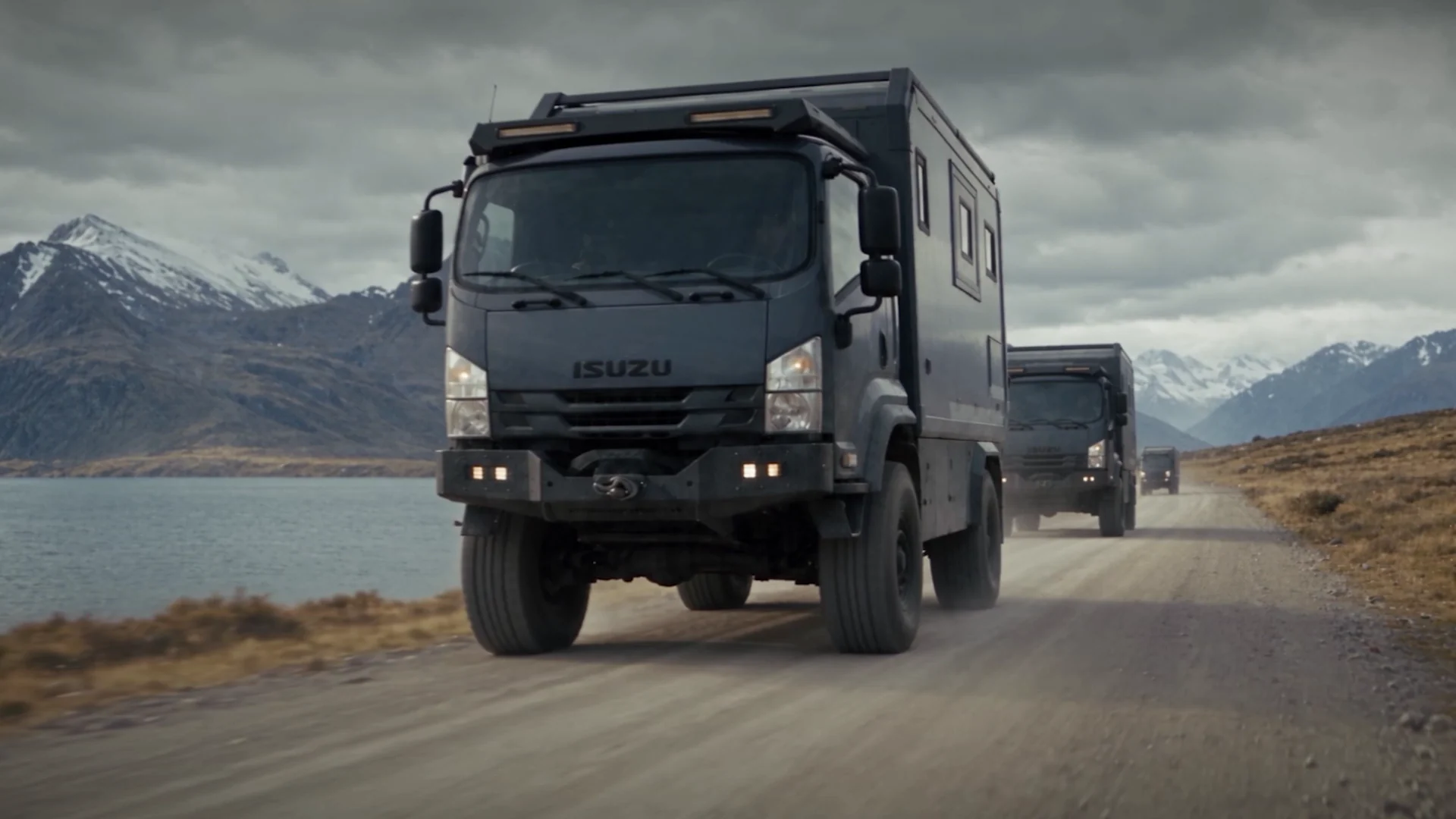 Three expedition trucks on a gravel road along a mountain lake