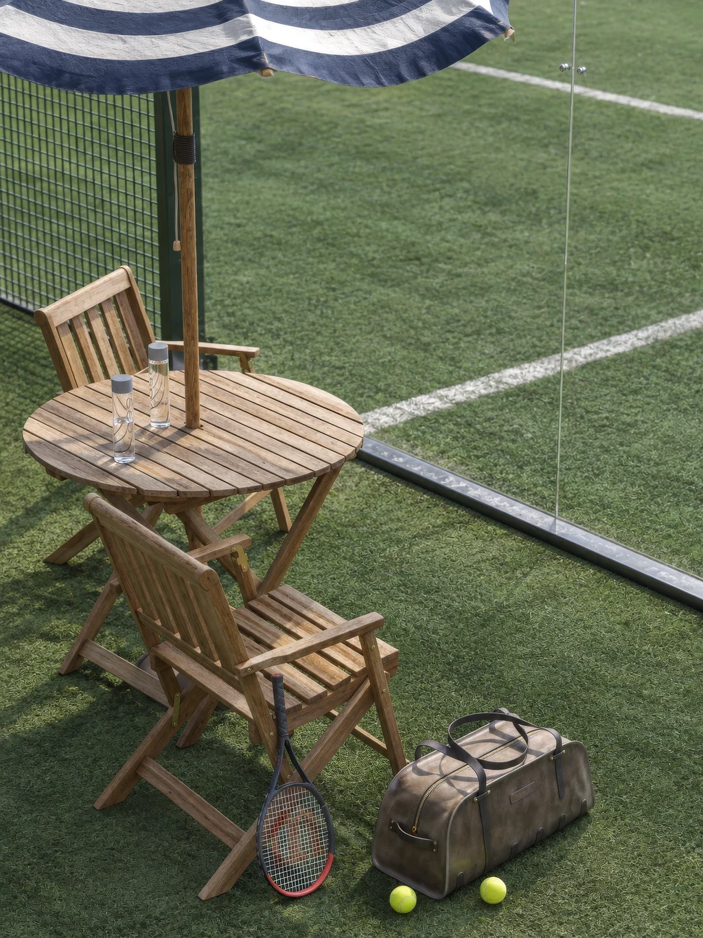 Render of a padel court rest area with a wooden table and chairs under a striped umbrella, a racket and a sports bag