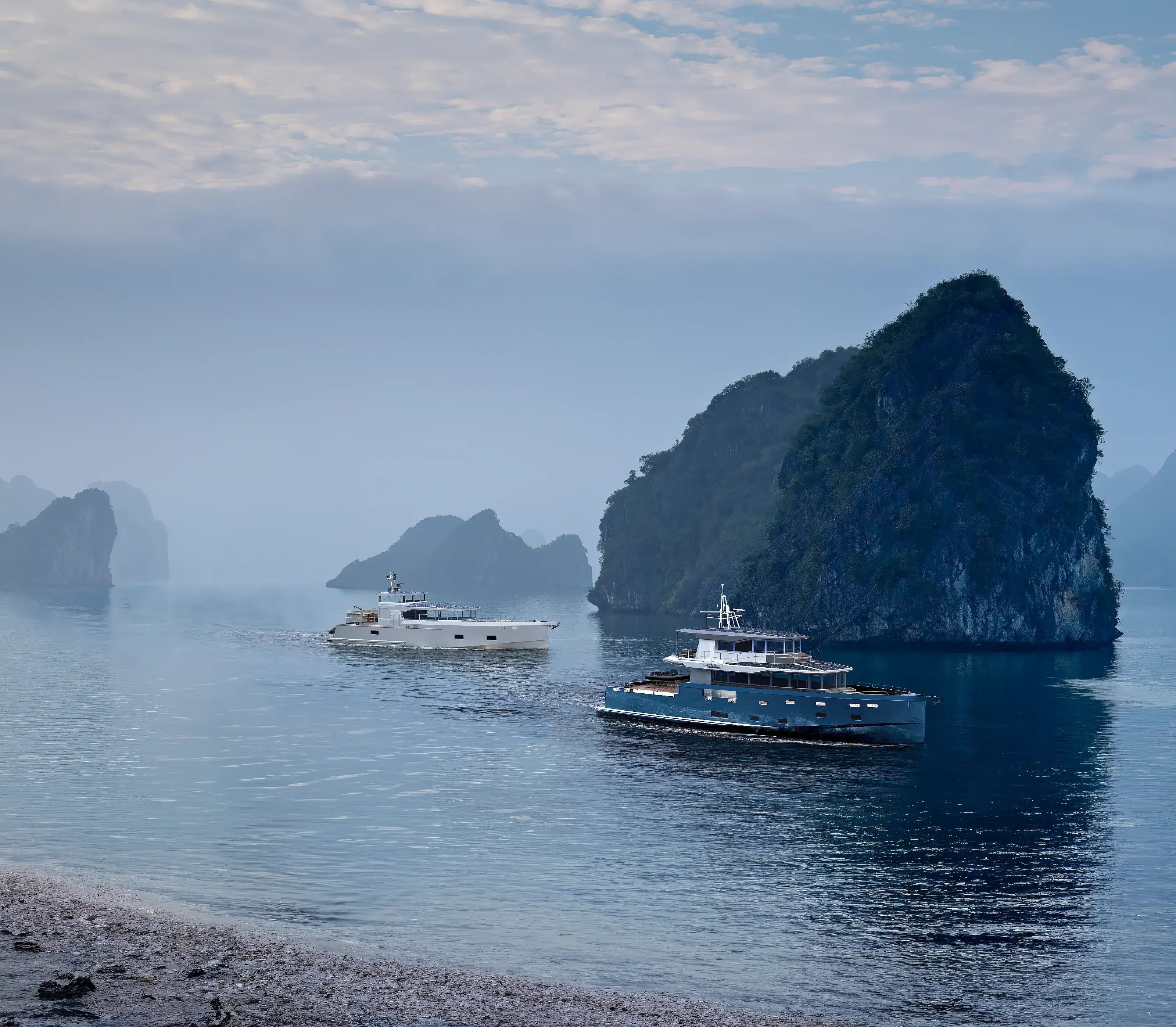 Two modern yachts moving through a calm bay surrounded by rocky islands and misty coastal landscape