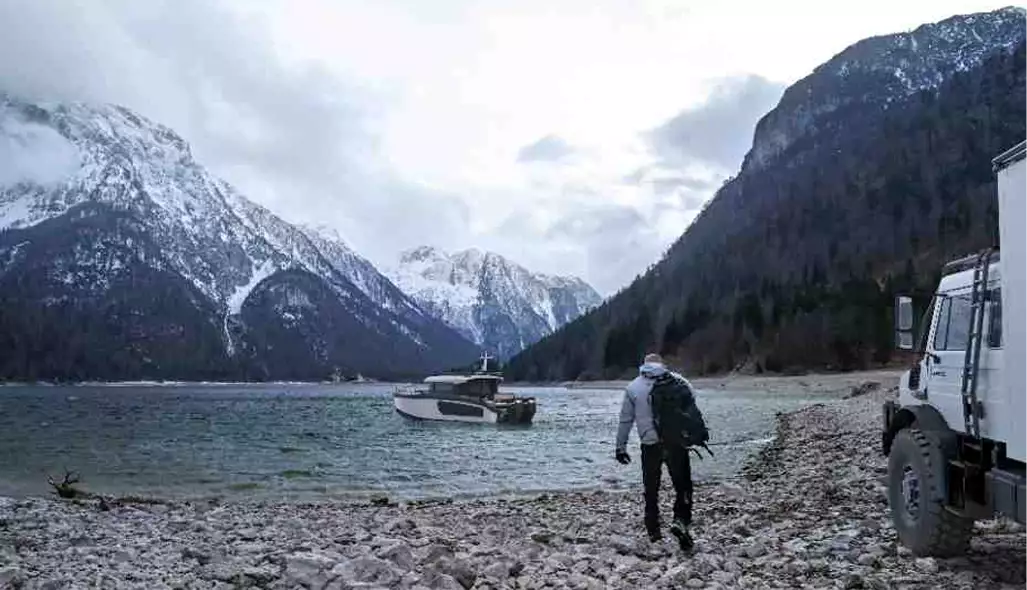 Boat renderings of a person walking toward a cabin boat on a rocky lakeshore below snowy mountains