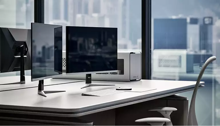 Desk close-up in skyscraper rendering with monitors, keyboard, chair, and city view through tall windows.