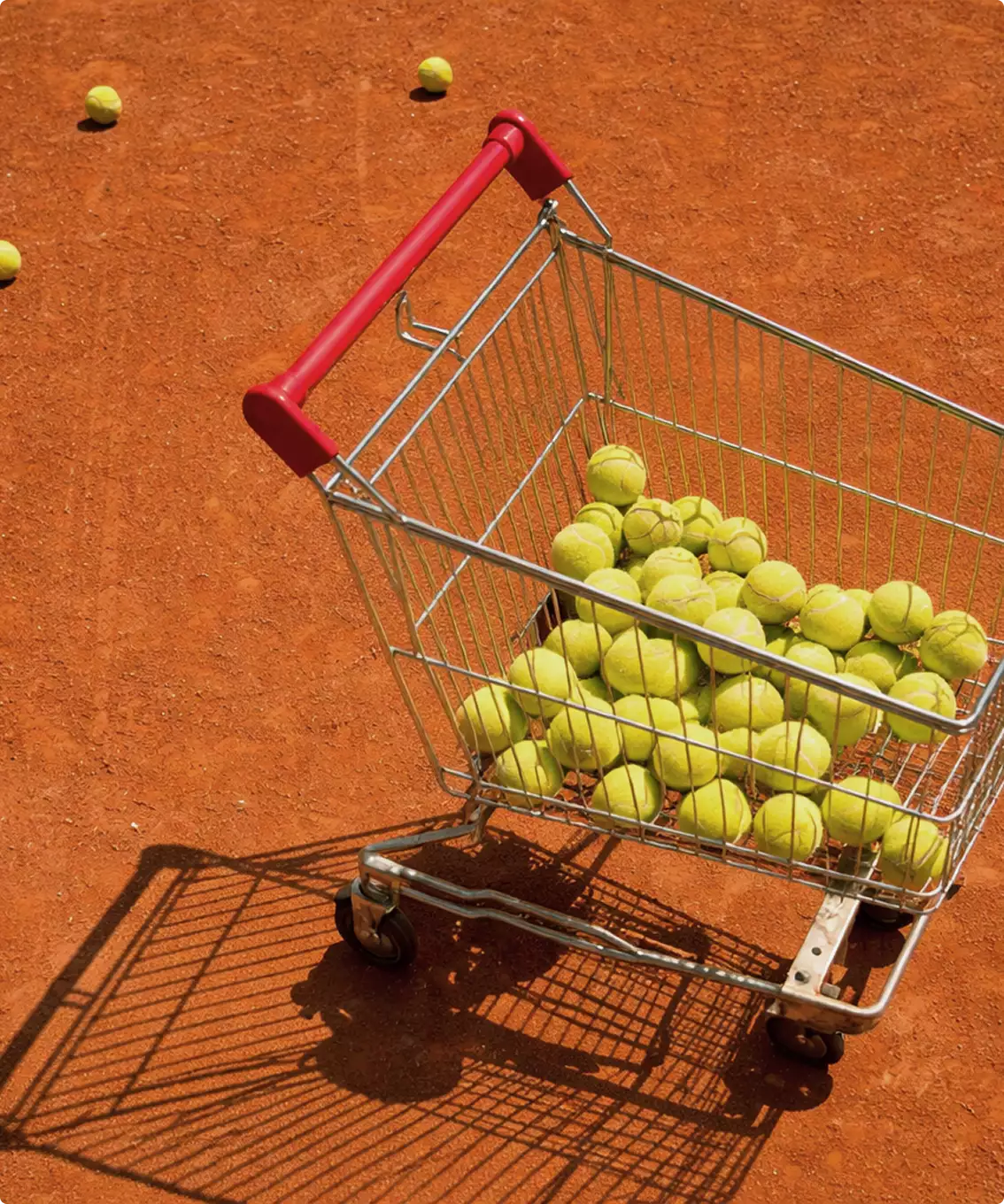 Shopping cart filled with tennis balls on clay tennis court design, scattered balls and shadow visible