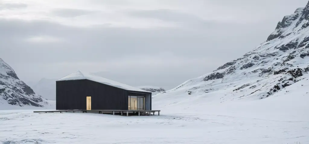 Winter rendering of a dark cabin in a snowy mountain valley with a lit doorway and overcast sky