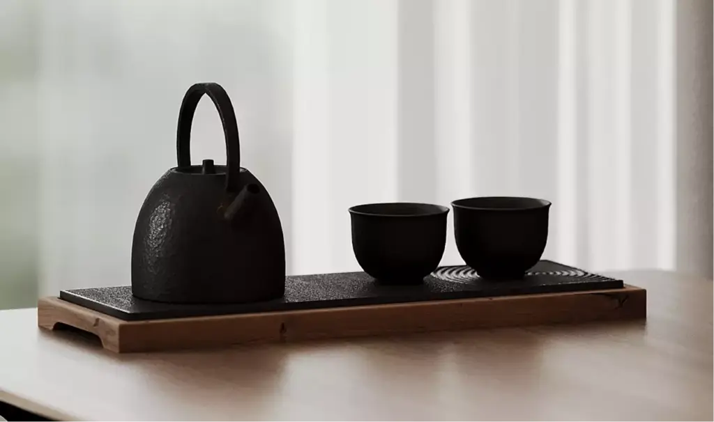 Tea set with kettle and cups on wooden tray near window light, black apartment design